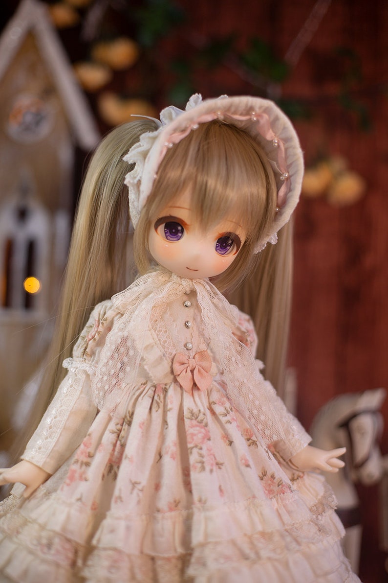 May include: A doll with long blonde hair styled in pigtails, wearing a white dress with pink floral accents and a white lace bonnet with pink trim.