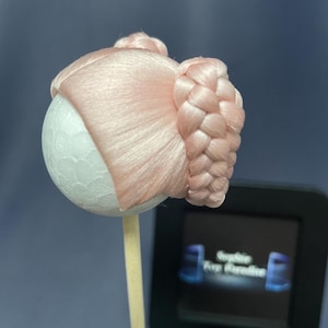 May include: A pink doll wig with two braided buns. The wig is on a styrofoam head and a wooden stick.