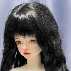 May include: A ball-jointed doll head featuring pale skin, light green eyes, and long, wavy black hair with bangs. The doll has a neutral facial expression with soft pink lips and subtle blush on the cheeks, showcasing detailed facial features.