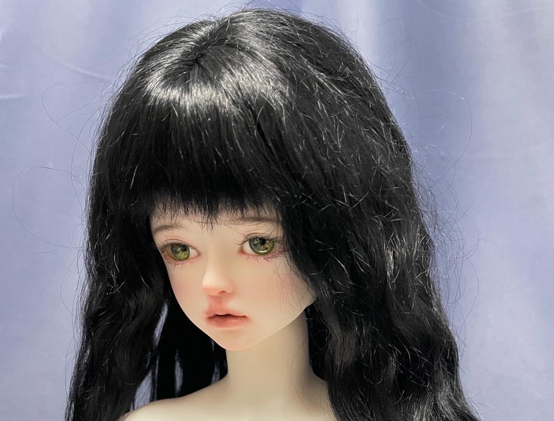 May include: A ball-jointed doll head featuring pale skin, light green eyes, and long, wavy black hair with bangs. The doll has a neutral facial expression with soft pink lips and subtle blush on the cheeks, showcasing detailed facial features.
