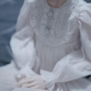 May include: A white, ruffled, long-sleeved dress with a button fastening at the neckline.