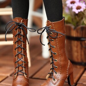 May include: Brown leather lace-up boots with a small heel. The boots have black laces and gold-coloured eyelets. The boots are on a wooden surface.