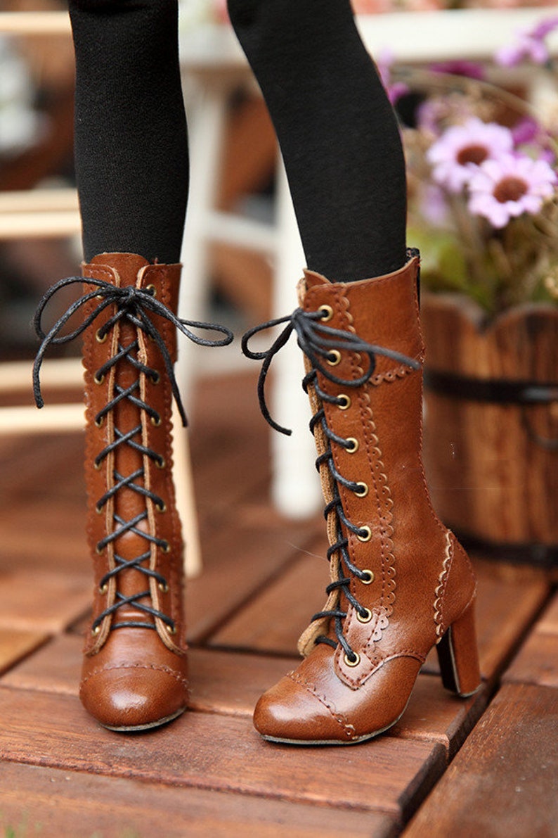 May include: Brown leather lace-up boots with a small heel. The boots have black laces and gold-coloured eyelets. The boots are on a wooden surface.