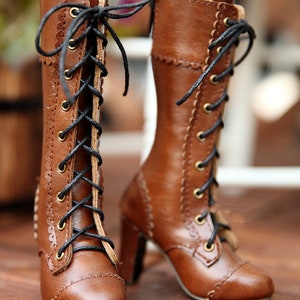 May include: A pair of brown leather lace-up boots with high heels. The boots have black laces, gold eyelets, and decorative stitching along the edges. The heels are a medium height, and the boots appear to be new.