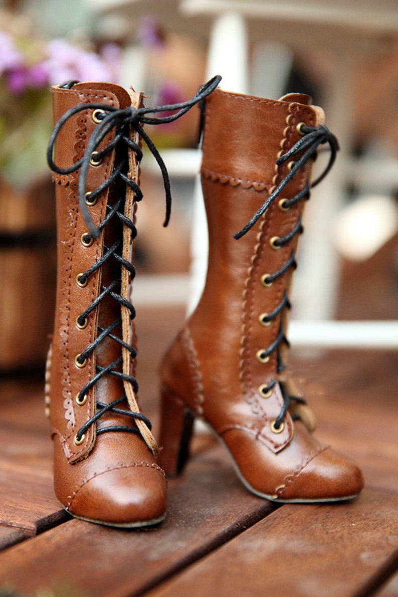 May include: A pair of brown leather lace-up boots with high heels. The boots have black laces, gold eyelets, and decorative stitching along the edges. The heels are a medium height, and the boots appear to be new.