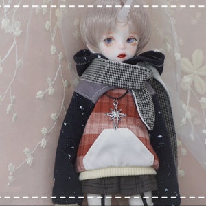 May include: A doll with short, silver hair wearing a red and black plaid hoodie with a white pocket, a black and white checkered scarf, and a silver star pendant necklace.