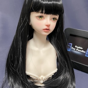May include: A doll head with long black hair and bangs. The doll has green eyes and a white porcelain face. The doll is wearing a white lace collar.