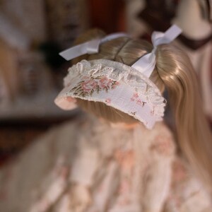 May include: A doll with long blonde hair wearing a white bonnet with pink floral embroidery and a white lace trim. The doll is wearing a white dress with a floral pattern.
