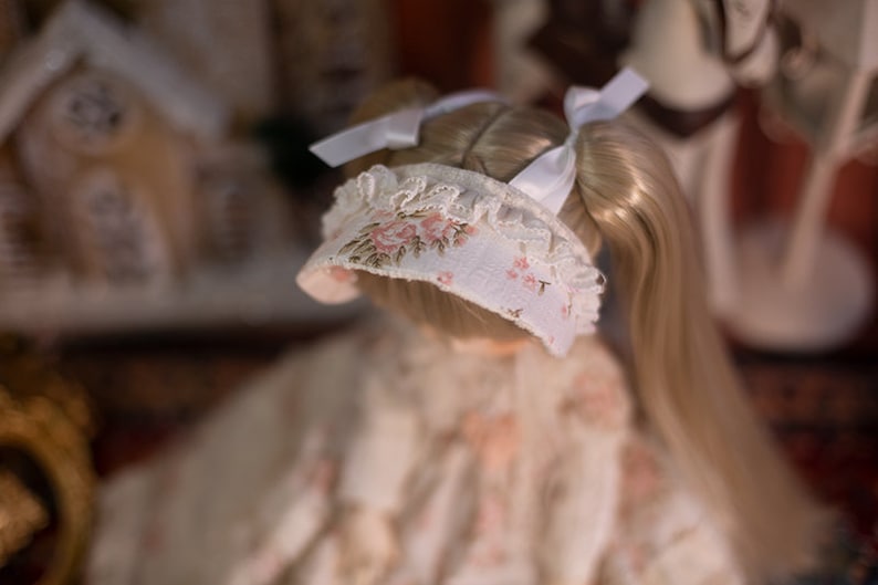 May include: A doll with long blonde hair wearing a white bonnet with pink floral embroidery and a white lace trim. The doll is wearing a white dress with a floral pattern.