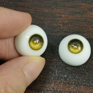 Puede incluir: Dos ojos de mu&ntilde;eca realistas con escler&oacute;tica blanca e iris dorado y negro. Los ojos est&aacute;n sobre una superficie de madera.