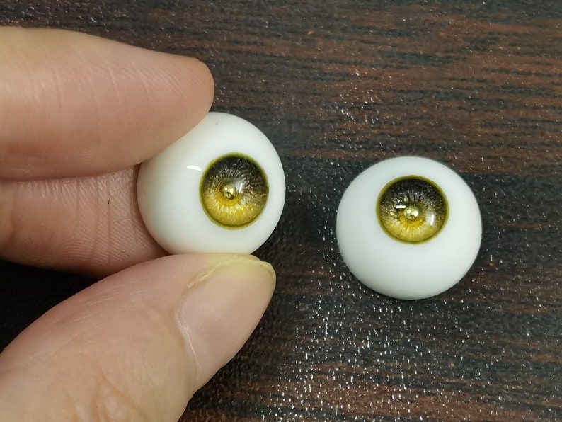Puede incluir: Dos ojos de mu&ntilde;eca realistas con escler&oacute;tica blanca e iris dorado y negro. Los ojos est&aacute;n sobre una superficie de madera.