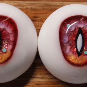 May include: A pair of realistic-looking glass eyes with a red, orange, and white iris. The eyes have a black pupil and a white sclera. The iris has a glittery texture and small blue and white flecks.