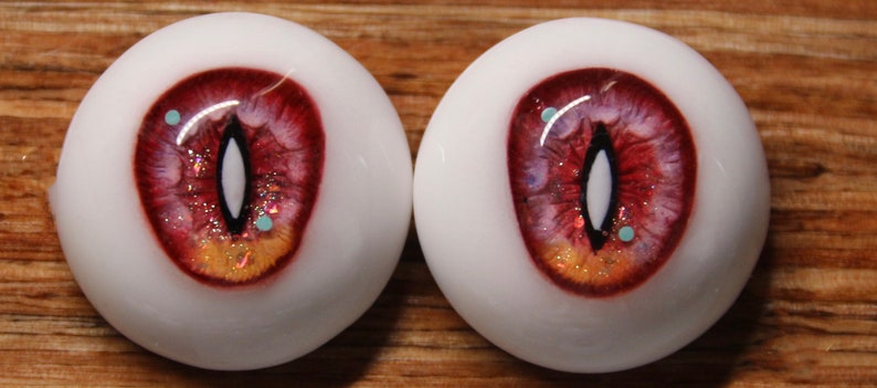 May include: A pair of realistic-looking glass eyes with a red, orange, and white iris. The eyes have a black pupil and a white sclera. The iris has a glittery texture and small blue and white flecks.