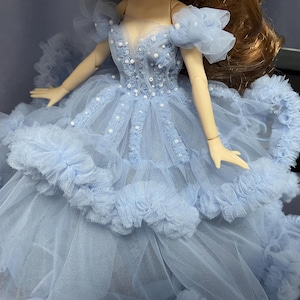 May include: A light blue tulle dress with a ruffled skirt and a beaded bodice. The dress is worn by a doll with long brown hair.