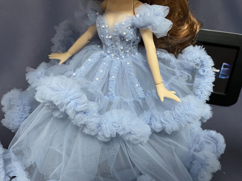 May include: A light blue tulle dress with a ruffled skirt and a beaded bodice. The dress is worn by a doll with long brown hair.