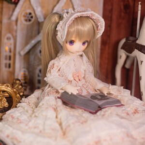May include: A doll wearing a white dress with pink accents and a white bonnet sits in front of a wooden toy house, holding a closed book. The doll has blonde hair and purple eyes.