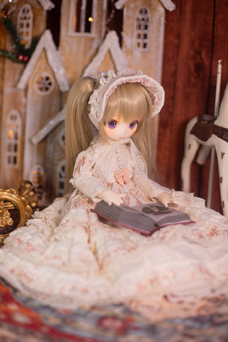May include: A doll wearing a white dress with pink accents and a white bonnet sits in front of a wooden toy house, holding a closed book. The doll has blonde hair and purple eyes.