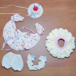 May include: A doll's clothing set featuring a floral print dress with blue ribbons, matching hat, bloomers, socks, and a large cream-colored ruffle. A small hat with a cherry on top is also included.