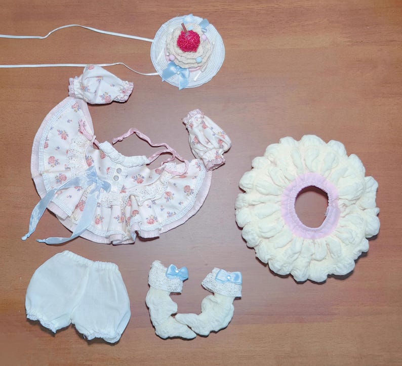 May include: A doll's clothing set featuring a floral print dress with blue ribbons, matching hat, bloomers, socks, and a large cream-colored ruffle. A small hat with a cherry on top is also included.