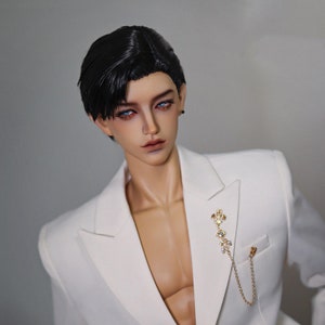 May include: A doll with short black hair wearing a white blazer with a gold chain and a decorative pin.