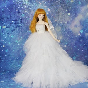 May include: A white tulle wedding dress with a strapless bodice and a long train. The dress is decorated with a white satin ribbon and a small rhinestone embellishment. The doll is wearing a white veil.