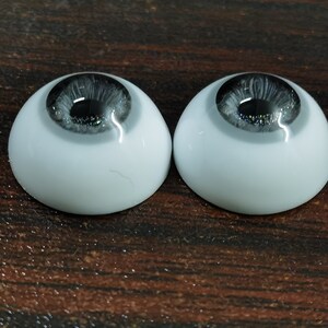 May include: Two gray and black doll eyes with a white sclera. The eyes are on a brown wooden surface.