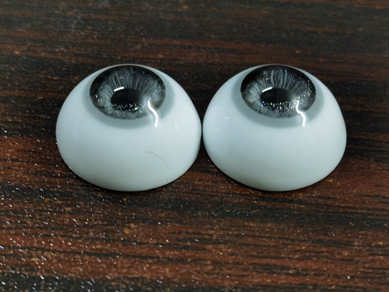 May include: Two gray and black doll eyes with a white sclera. The eyes are on a brown wooden surface.