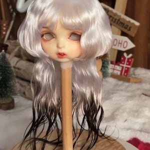 May include: A doll head with a pale complexion, white hair, and dark brown tips. The doll's face has a neutral expression with red lips and eye sockets. The head is mounted on a wooden stand, suggesting a craft or collectible item.