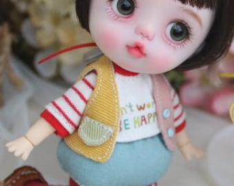 May include: A small doll with brown hair, wearing a yellow vest with a pocket, a white shirt with the text "Don't worry be happy", red and white striped sleeves, blue pants, and red and white polka dot tights.
