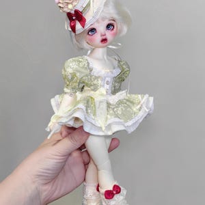 May include: A doll with white hair, blue eyes, and rosy cheeks, wearing a green dress with white lace trim. The doll has a matching hat with a cherry on top and is wearing white shoes with a small heel. The doll is being held by a hand.