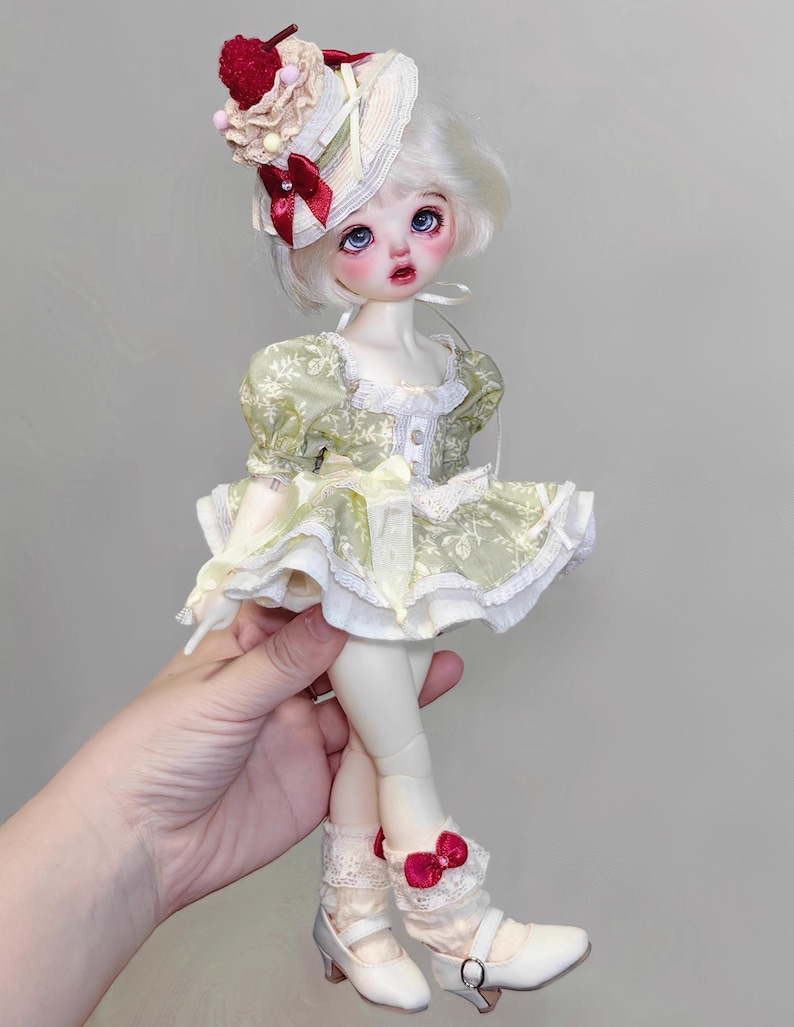 May include: A doll with white hair, blue eyes, and rosy cheeks, wearing a green dress with white lace trim. The doll has a matching hat with a cherry on top and is wearing white shoes with a small heel. The doll is being held by a hand.