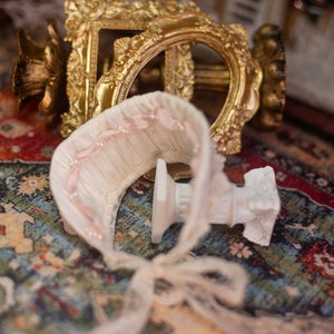 May include: A white lace and pink ribbon baby bonnet with a white pedestal base, sitting on a colorful rug with a gold ornate frame in the background.
