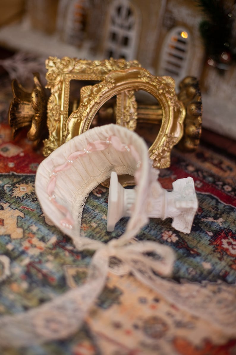 May include: A white lace and pink ribbon baby bonnet with a white pedestal base, sitting on a colorful rug with a gold ornate frame in the background.