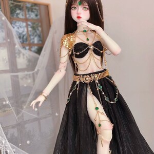 May include: A doll wearing a black and gold costume with green gem accents. The costume includes a halter top, a skirt, and a headpiece. The doll has long brown hair and green eyes.