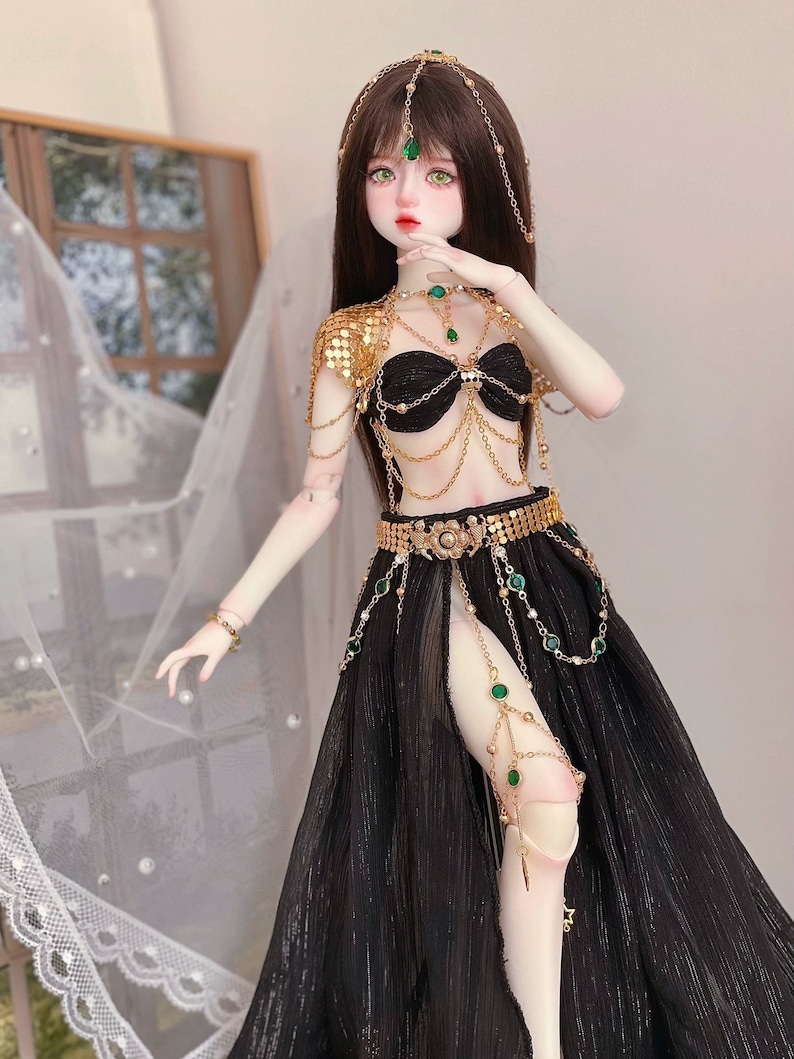 May include: A doll wearing a black and gold costume with green gem accents. The costume includes a halter top, a skirt, and a headpiece. The doll has long brown hair and green eyes.