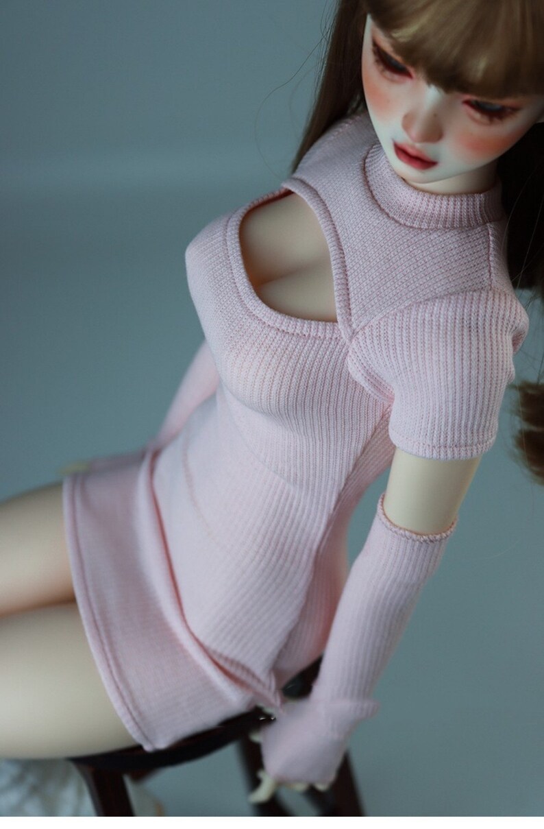 May include: A pink knitted doll dress with a cut-out detail at the neckline and long sleeves. The dress has a fitted silhouette and a short skirt.