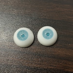 K&ouml;nnte beinhalten: Zwei wei&szlig;e Puppenaugen mit blauen Iris. Die Augen sind rund und haben ein realistisches Aussehen.