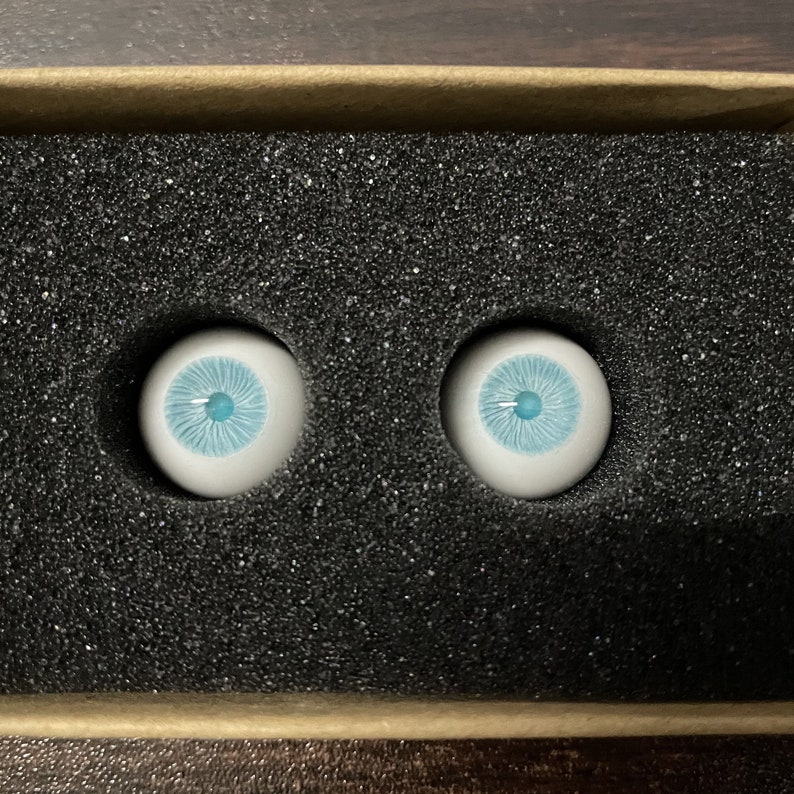K&ouml;nnte beinhalten: Zwei blaue Glasaugen mit wei&szlig;en Iris und blauen Pupillen, in einer schwarzen, mit Schaumstoff ausgekleideten Box.