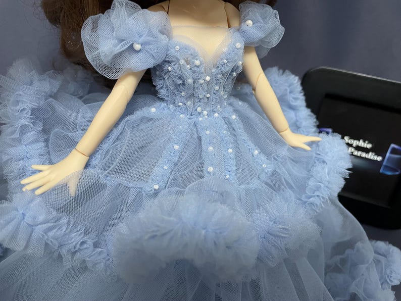 May include: A light blue tulle dress with a sweetheart neckline and pearl embellishments. The dress has a ruffled skirt and a large flower on each shoulder.