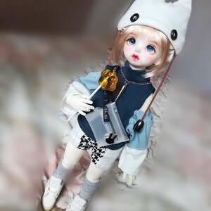 May include: A doll with blonde hair wearing a white cat ear hat, a blue and white striped shirt, and white shorts. The doll is holding a gold lollipop and a clear plastic bag.