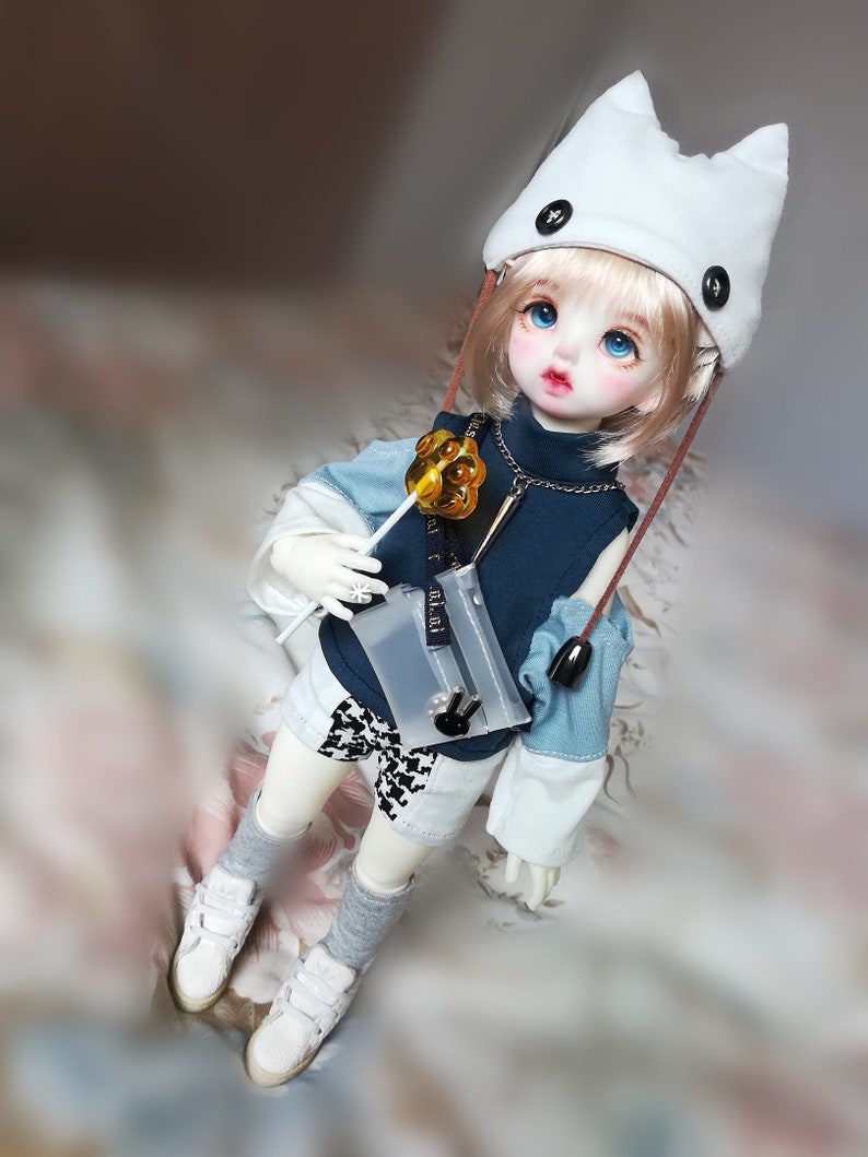 May include: A doll with blonde hair wearing a white cat ear hat, a blue and white striped shirt, and white shorts. The doll is holding a gold lollipop and a clear plastic bag.
