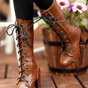 May include: Brown leather lace-up boots with a high heel. The boots have a scalloped edge design and black laces. The boots are worn with black leggings. The background includes a wooden barrel and flowers.