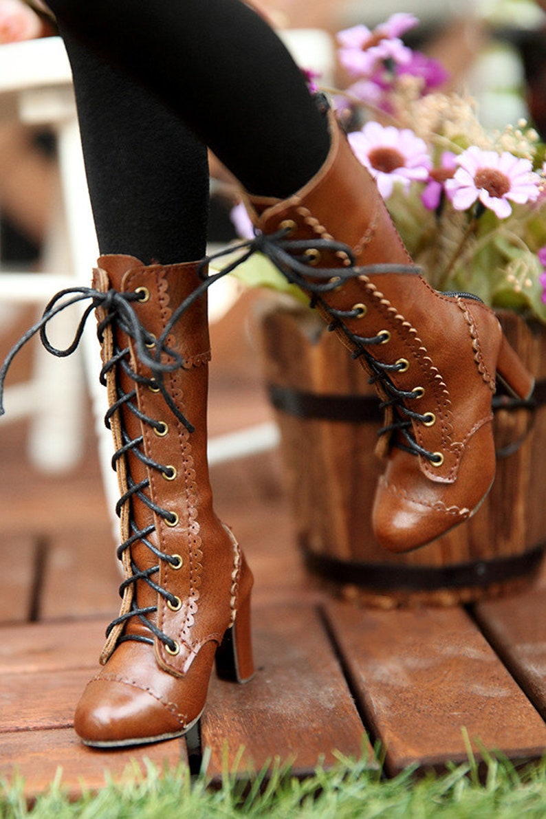 May include: Brown leather lace-up boots with a high heel. The boots have a scalloped edge design and black laces. The boots are worn with black leggings. The background includes a wooden barrel and flowers.