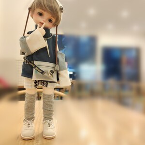 May include: A doll with blonde hair wearing a blue and white outfit with a brown belt and a white headband. The doll is standing on a wooden surface.