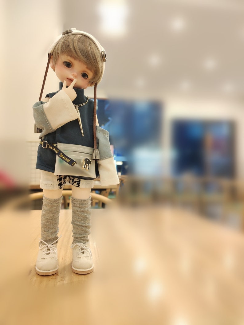 May include: A doll with blonde hair wearing a blue and white outfit with a brown belt and a white headband. The doll is standing on a wooden surface.