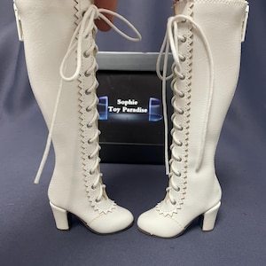 Pode incluir: Um par de botas brancas, at&eacute; ao joelho, com atacadores e saltos pequenos. As botas t&ecirc;m uma guarni&ccedil;&atilde;o decorativa nas laterais e um detalhe de renda com babados na parte superior. O texto "Sophie Toy Paradise" &eacute; vis&iacute;vel numa caixa no fundo.
