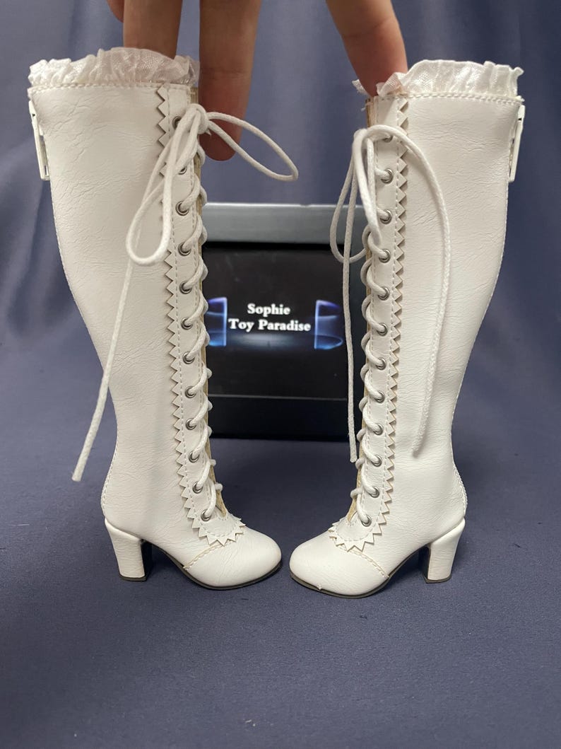 Pode incluir: Um par de botas brancas, at&eacute; ao joelho, com atacadores e saltos pequenos. As botas t&ecirc;m uma guarni&ccedil;&atilde;o decorativa nas laterais e um detalhe de renda com babados na parte superior. O texto "Sophie Toy Paradise" &eacute; vis&iacute;vel numa caixa no fundo.