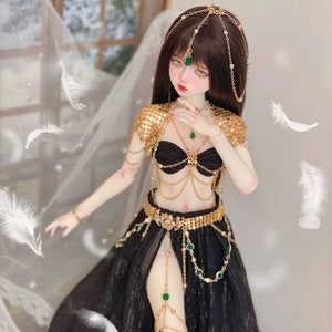 May include: A doll wearing a black and gold costume with green gem accents. The doll has long brown hair and is wearing a gold headpiece with green gems. The doll is posed in a dancing position.