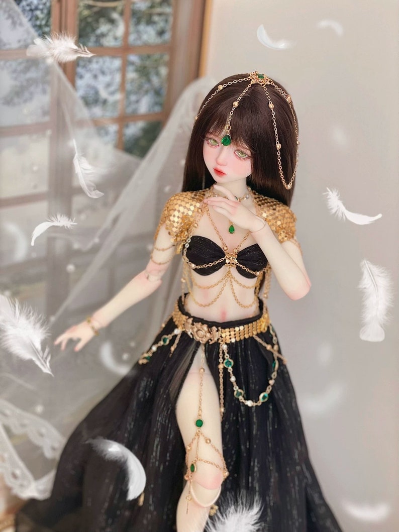 May include: A doll wearing a black and gold costume with green gem accents. The doll has long brown hair and is wearing a gold headpiece with green gems. The doll is posed in a dancing position.