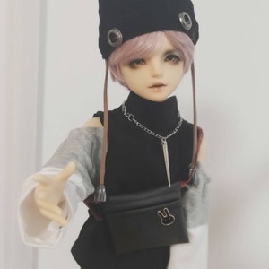 May include: A doll with pink hair wearing a black hat with ear flaps, a black turtleneck, a gray and white long-sleeved shirt, black shorts with a white and black checkered pattern, and a black fanny pack with a bunny design.